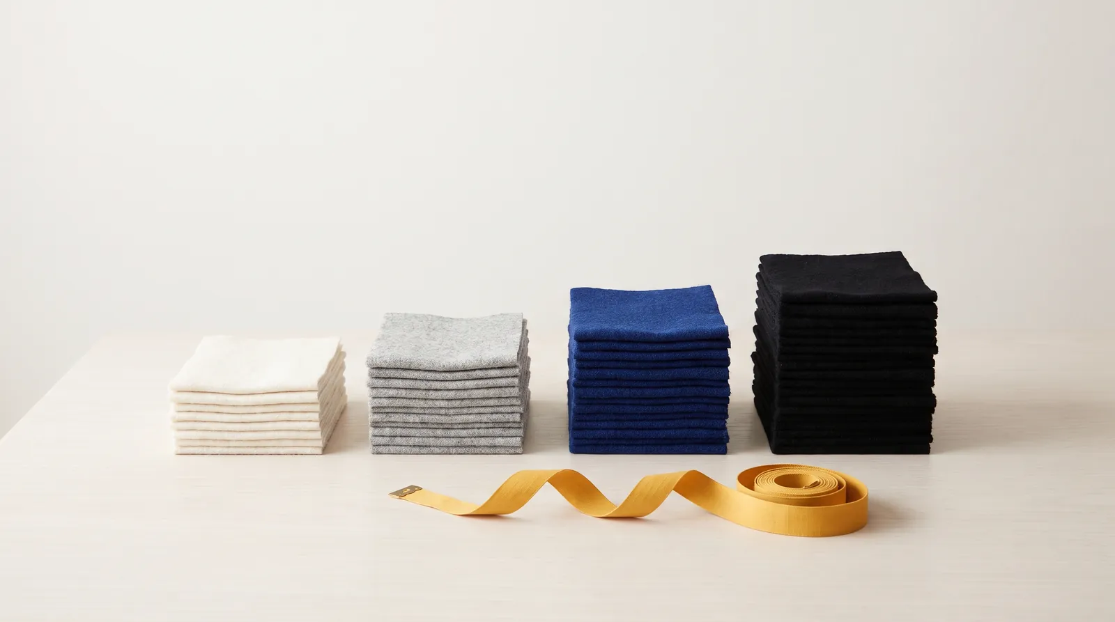 Fabric samples in white, grey, cobalt blue and black, layered into four stacks of increasing height, with a yellow tape measure in front